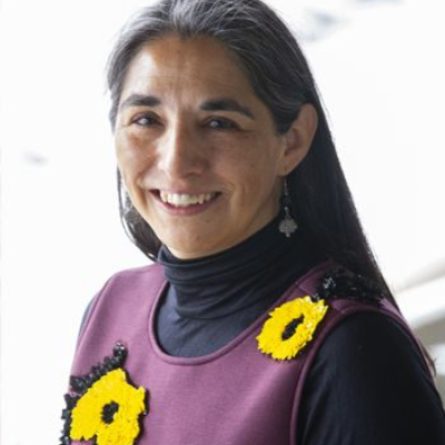 Portrait of Ingrid Aguayo Fuentealba wearing a purple sleeveless dress over a black turtleneck, with two yellow flower pins, facing the camera against a softly blurred background.